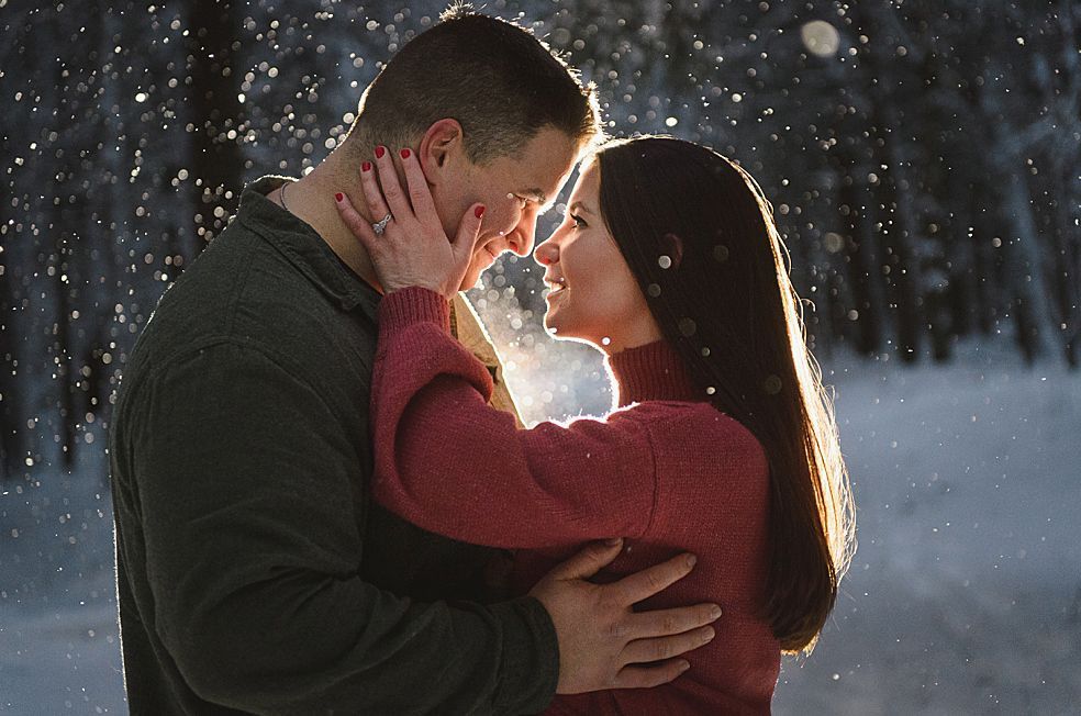Couple embracing in snowy forest, backlit by sun. Woman in red sweater, man in green, smiling.