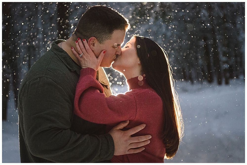 Couple kissing in a snowy forest, with a bright sunlit backdrop.
