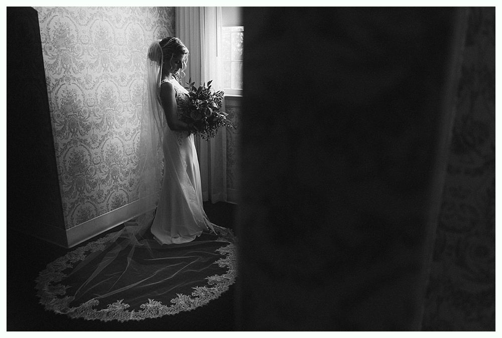 Bride in wedding dress, holding bouquet, leans against patterned wall near a window. Black and white photo.
