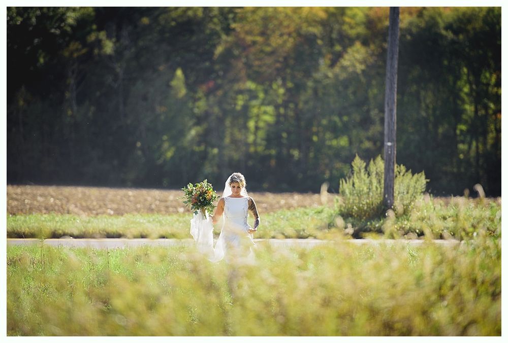 Bride in white dress walking down a path, holding flowers, with a field and trees in the background.