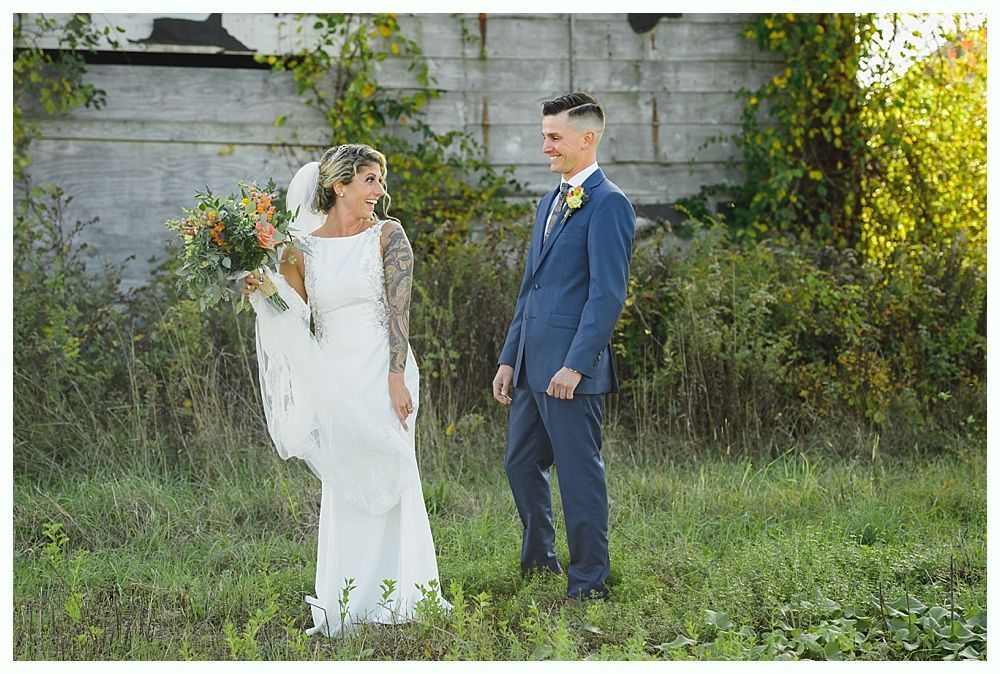 Bride and groom smile, outdoors, in front of a rustic building. Bride holds bouquet, groom in blue suit.