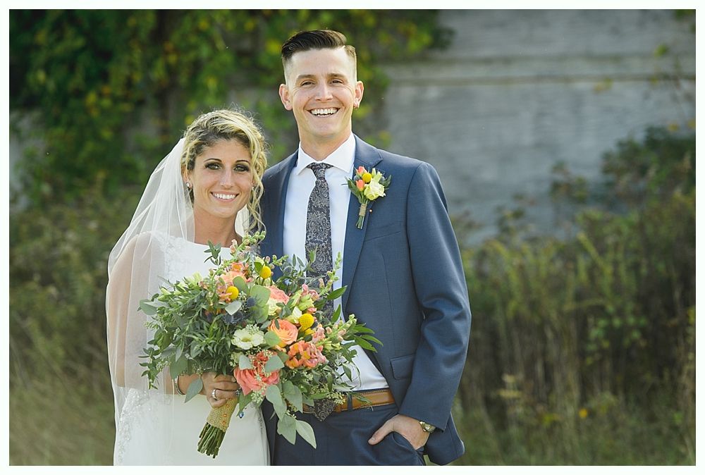 Bride and groom smiling at camera, bride holds bouquet, man in blue suit. Outdoor setting.