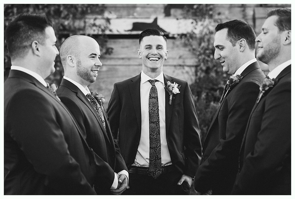 Groom smiling, surrounded by groomsmen in suits, outdoor setting, black and white photo.