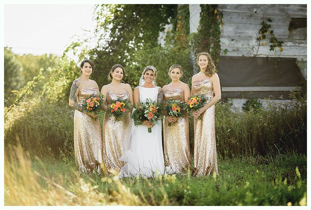 Bride and bridesmaids in gold sequin gowns holding bouquets posing outdoors by an old building.