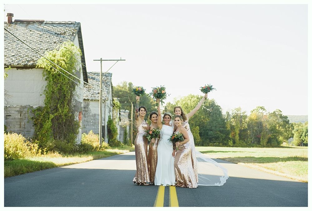 Bride and bridesmaids in gold gowns raise bouquets on a road near a weathered building.