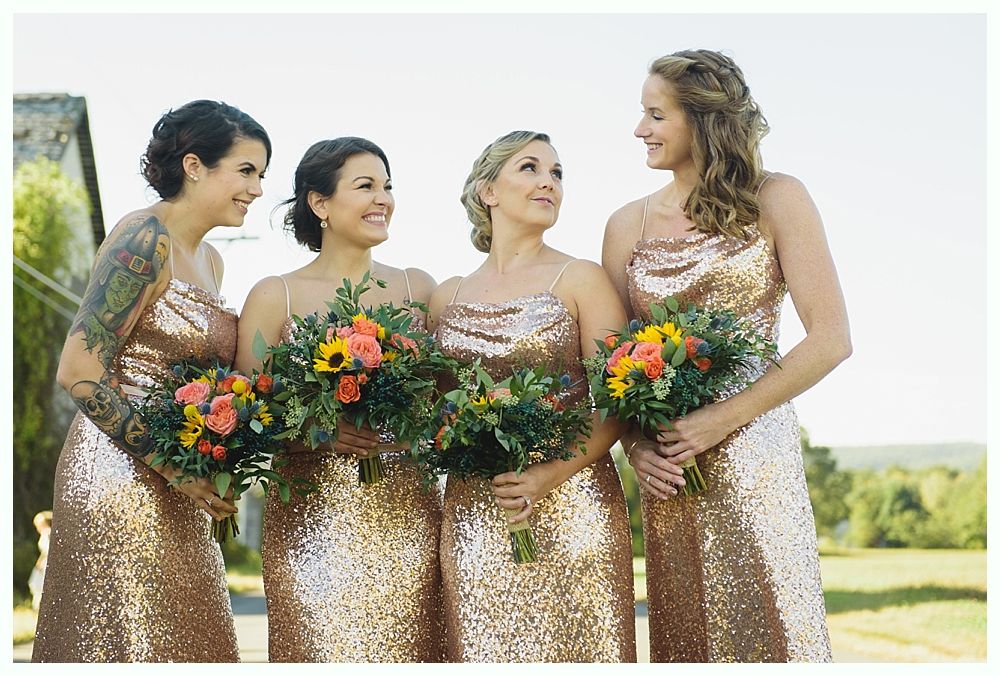 Four women in gold sequined gowns hold bouquets, smiling in a field.
