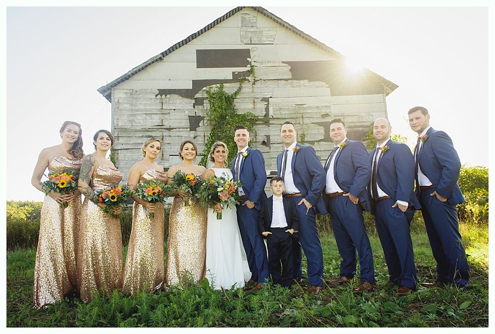 Wedding party poses in front of an old barn; gold sequined bridesmaid dresses, blue suits, sunny outdoor setting.