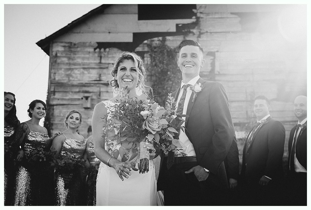Bride and groom smiling outdoors with wedding party; sunlit, old building in background. Black and white.