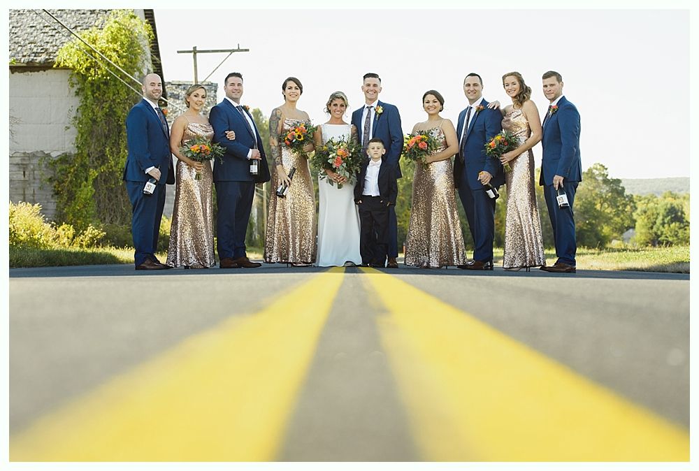 Wedding party stands in road. Bride and bridesmaids in gold, groomsmen in blue suits.