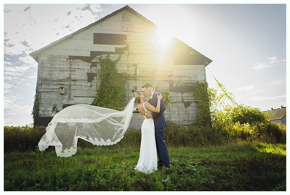 Couple kissing in front of a weathered barn, veil blowing in the wind, bright sunlight.