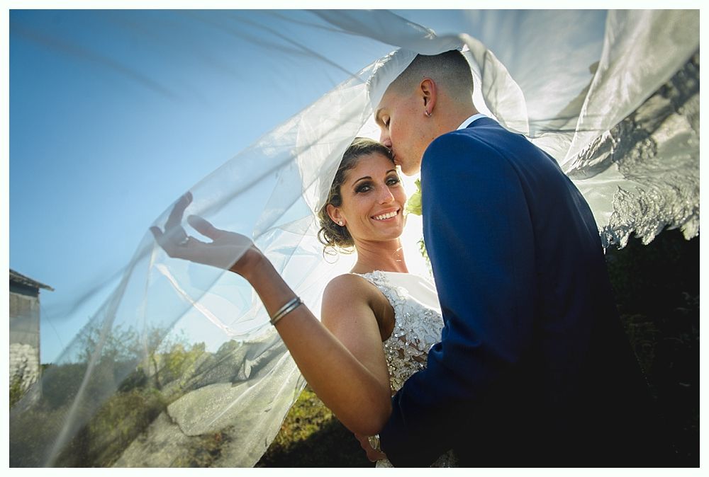 A couple embraces under a veil on a sunny day. The man kisses the woman's forehead as she smiles.