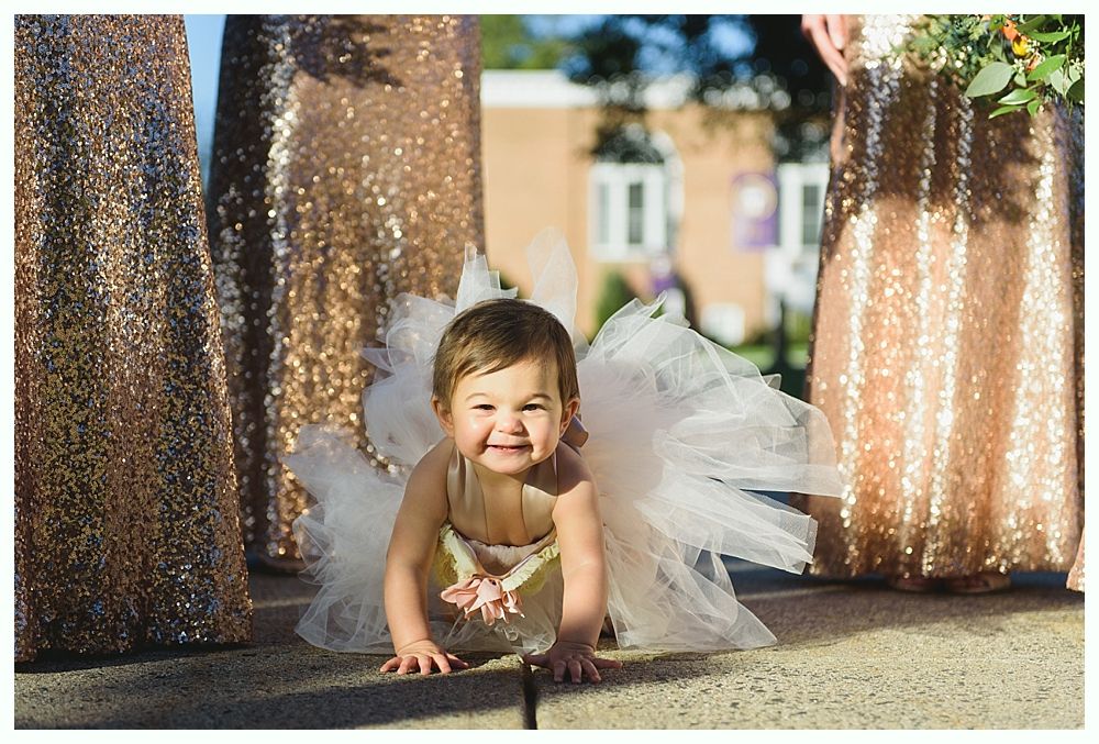 Smiling baby crawling on a sidewalk, wearing a white tutu, with bridesmaids in sequined dresses.