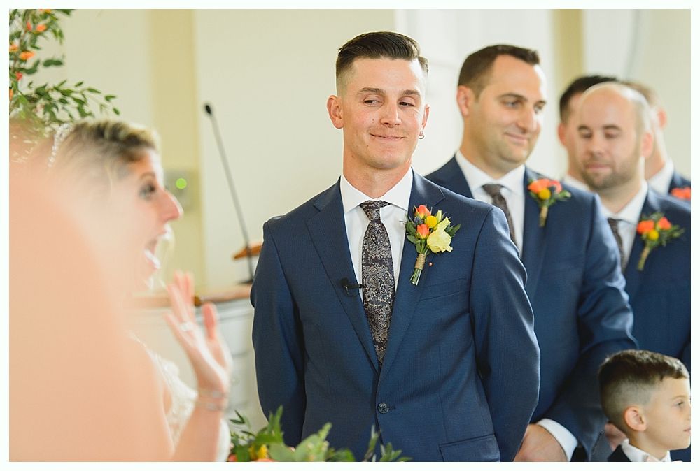 Groom smiling at bride during wedding ceremony; groomsmen in navy suits stand beside him.