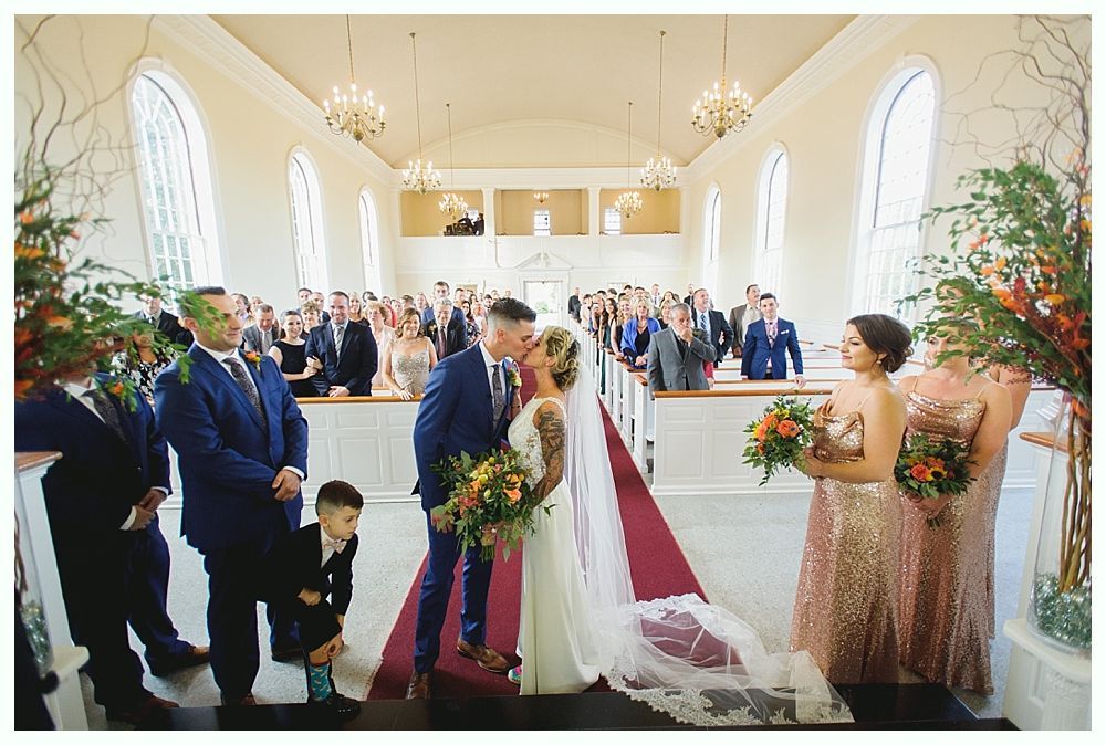 Bride and groom kissing at altar; wedding guests watch. Red carpet leads to arch window.