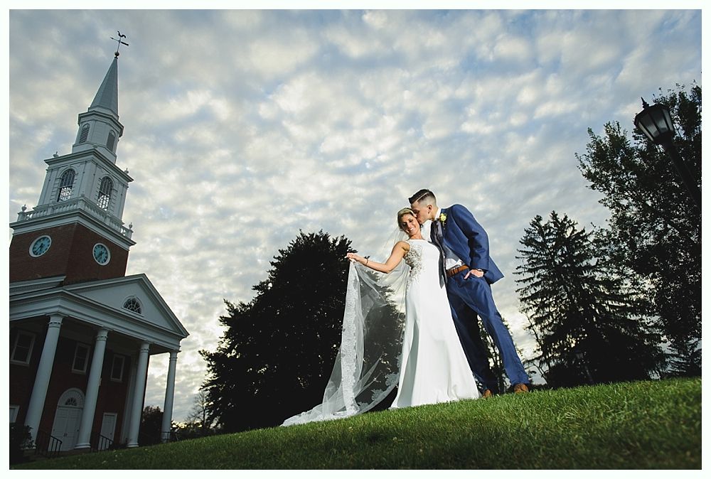 Bride and groom kissing, posed on a grassy hill with a church and cloudy sky in the background.
