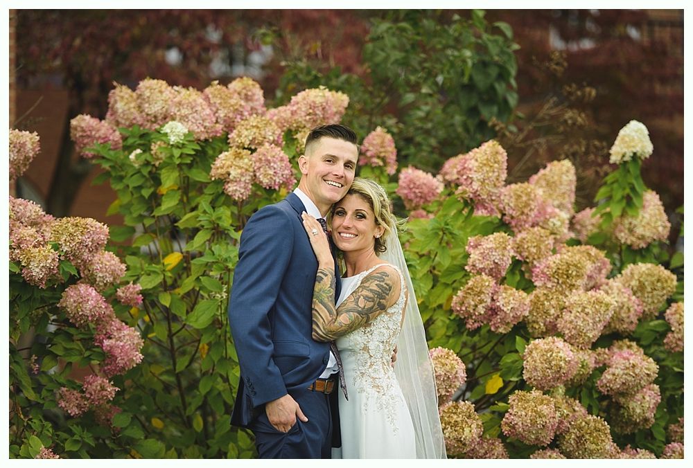 Newlyweds smiling in front of pink hydrangea bushes. Man in blue suit, woman in white wedding dress with arm tattoo.