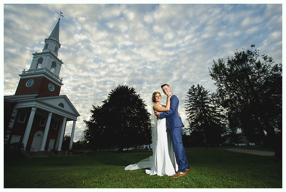 Couple embraces on grass near a church with a spire; cloudy sky.