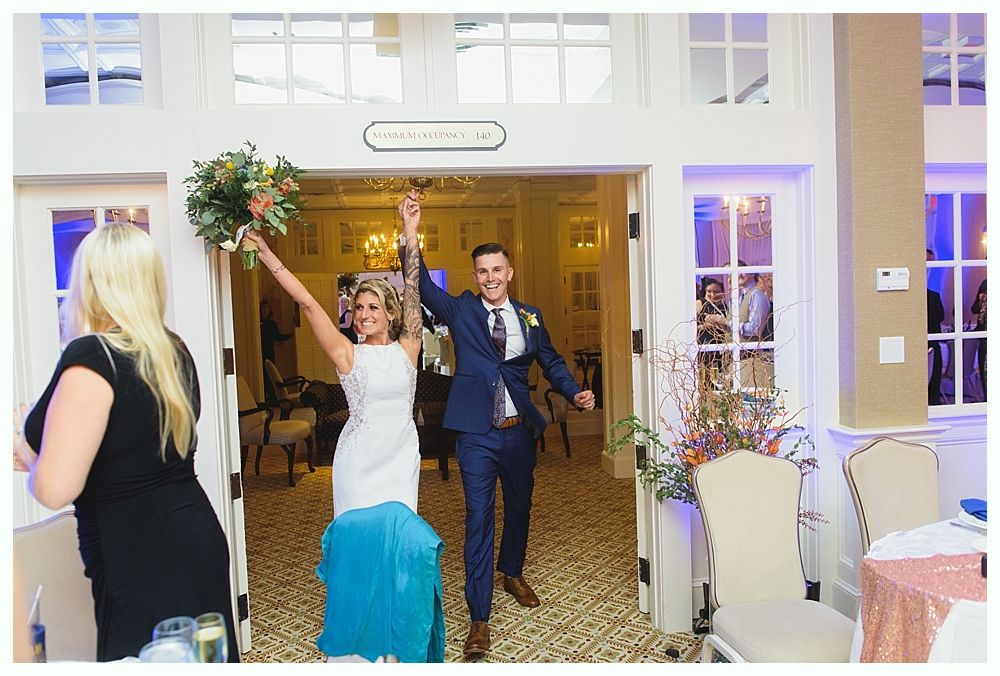 Bride and groom entering a reception, arms raised, smiling. Room with tables and windows.