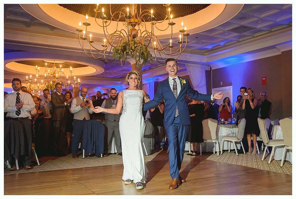 Newlyweds walk into reception, arms outstretched, smiling. Guests clap. Ballroom with chandelier.
