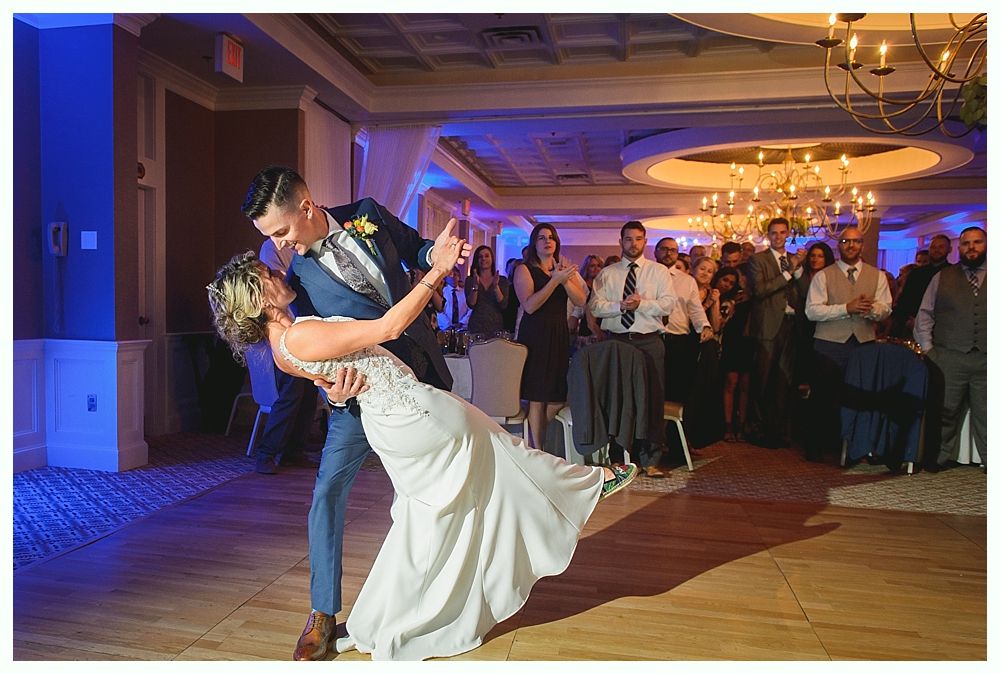 Bride and groom dance at reception; she leans back, he supports her; guests watch and applaud.
