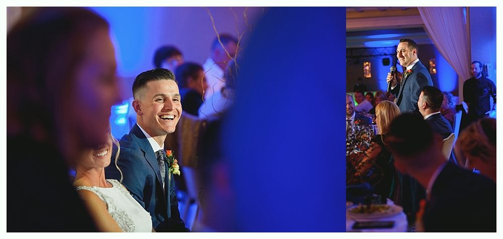 Guests at a wedding, one man smiling at the left, another man giving a speech in a room with blue lighting.