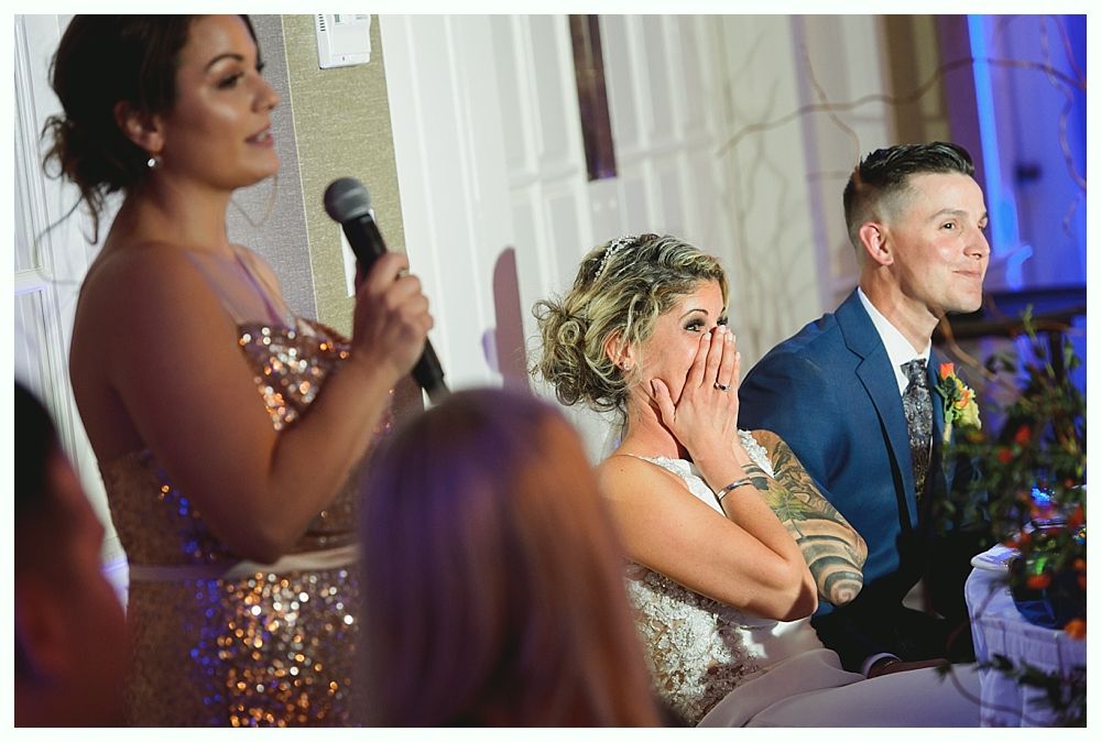 Woman giving speech at wedding, bride covers face, groom listens.