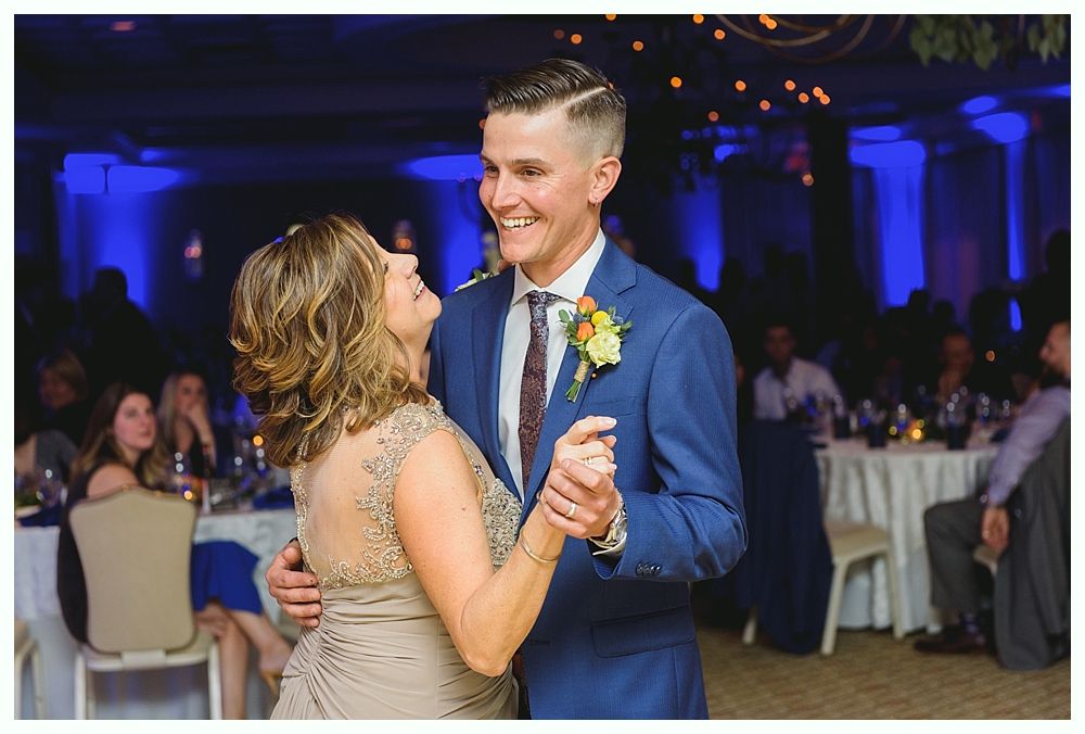 Man in blue suit dances with woman in beige dress at an event. They smile, facing each other. Blue-lit room.