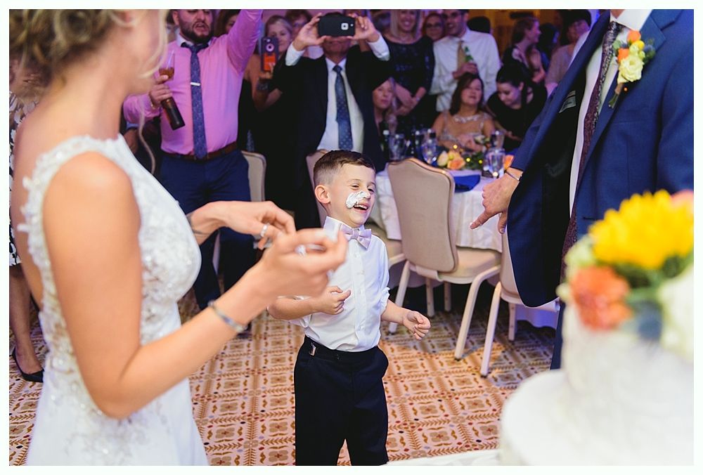 Child with frosting on face at a wedding, being fed cake by a bride and groom.