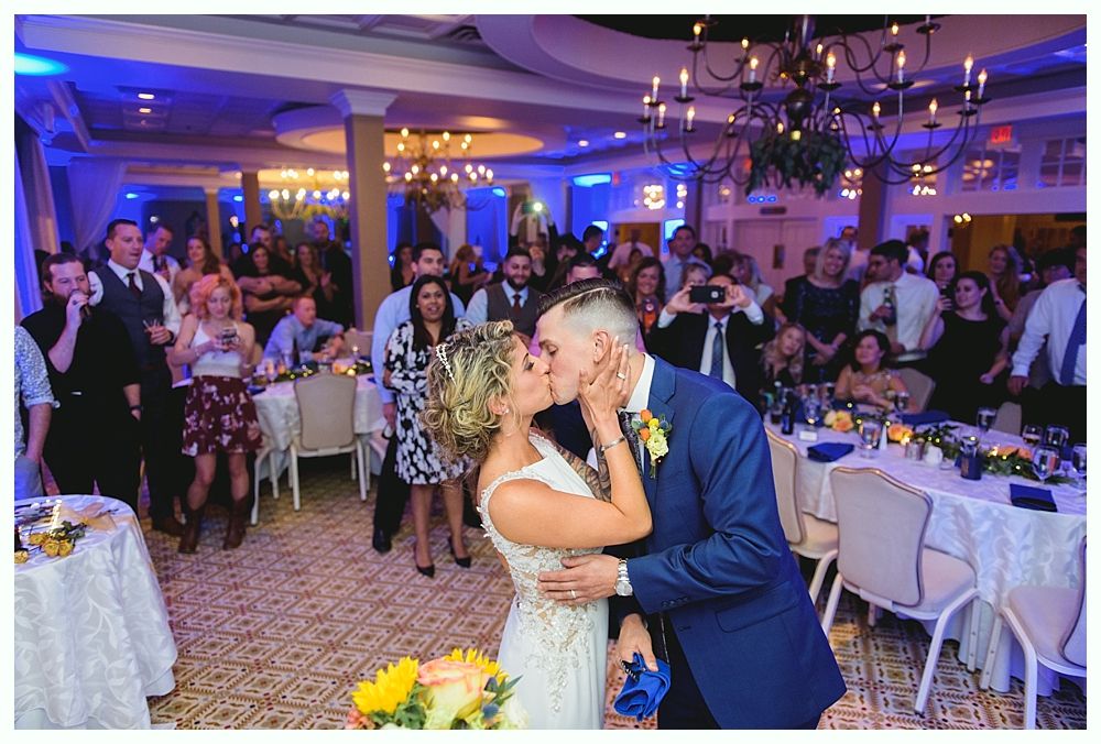 Bride and groom kissing, surrounded by guests at a wedding reception with tables, and a chandelier in a decorated ballroom.