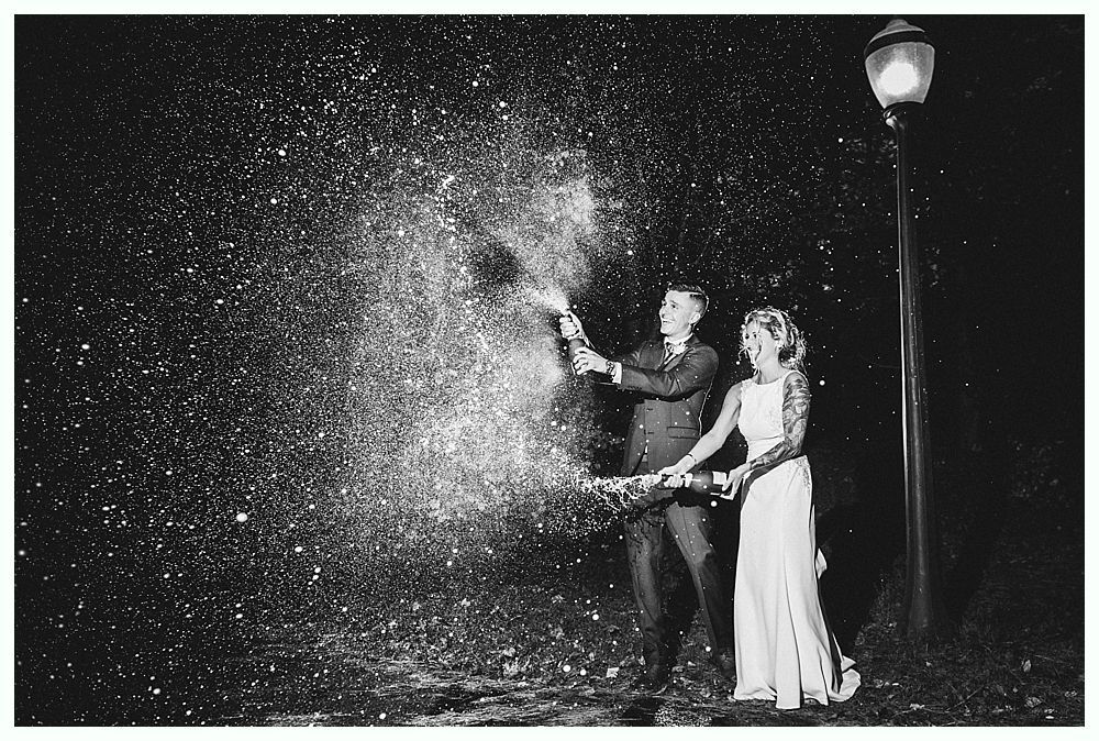 Newlyweds pop champagne, spraying sparkles in the night. A street lamp stands nearby.