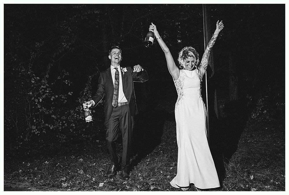 Newlyweds celebrate with champagne bottles, arms raised, in dark outdoor setting.