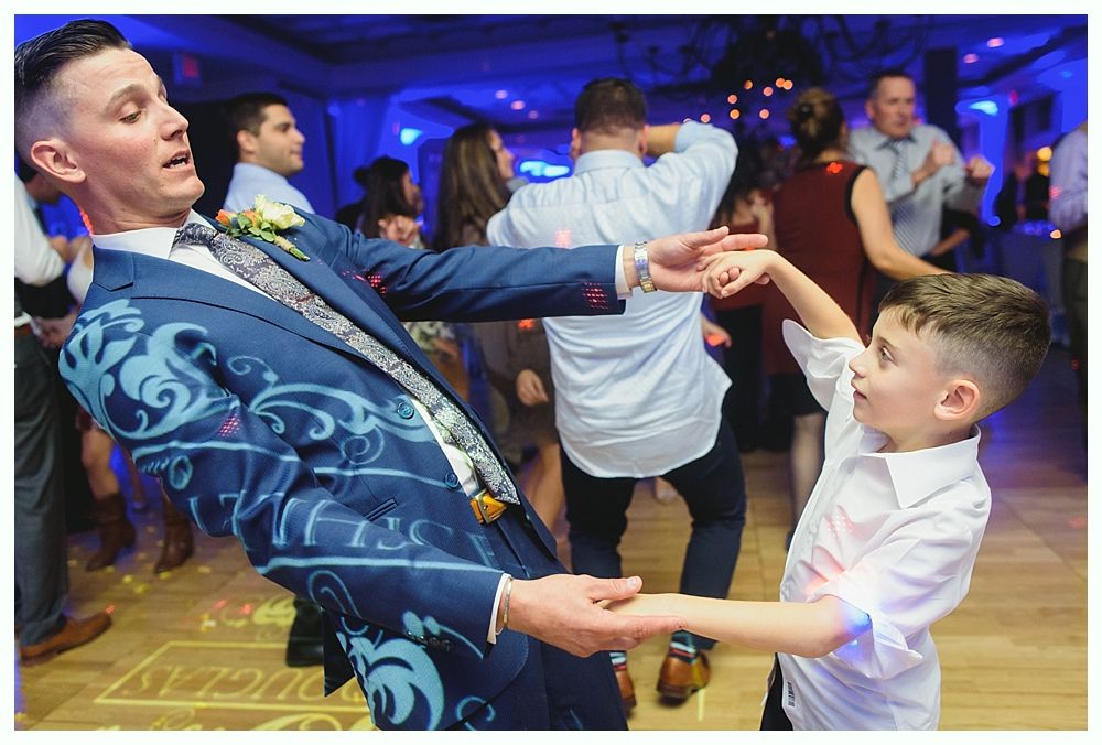 Man in patterned suit dances with a child on a lit dance floor. Other people dance in the background.