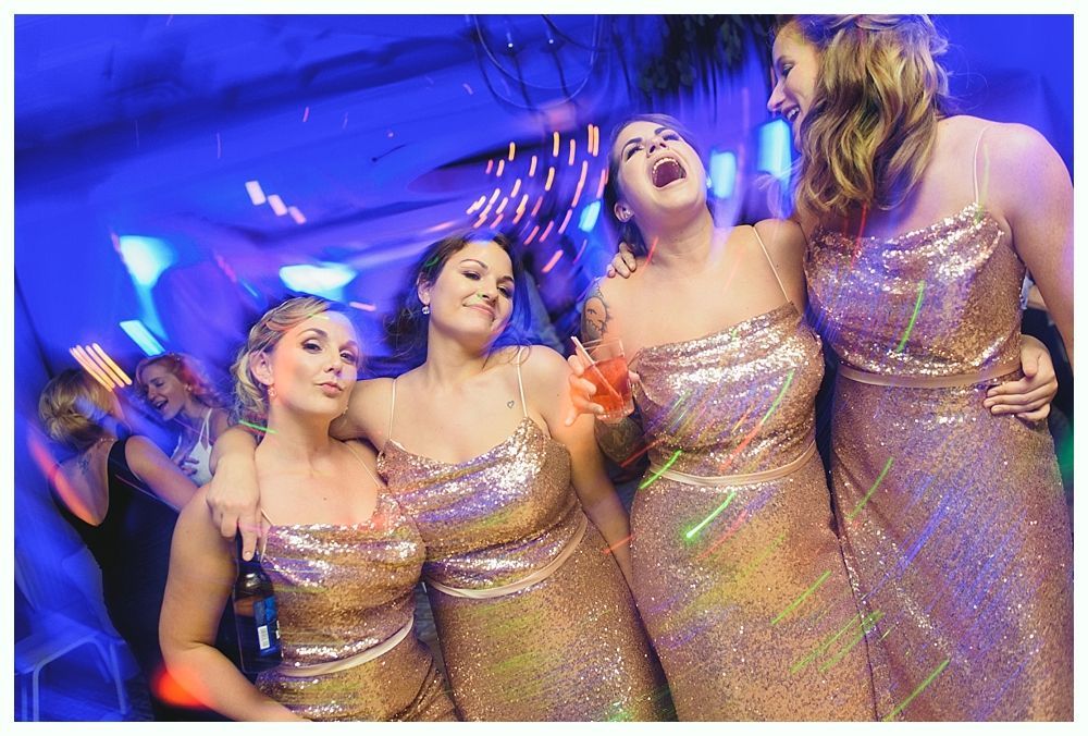 Four women in gold sequin dresses, laughing and hugging, at a party with blue lighting.
