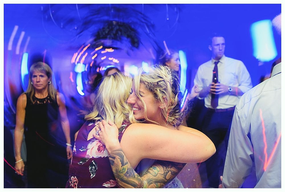 Two women embrace at a party with a swirling blue light effect, other people are in the background.