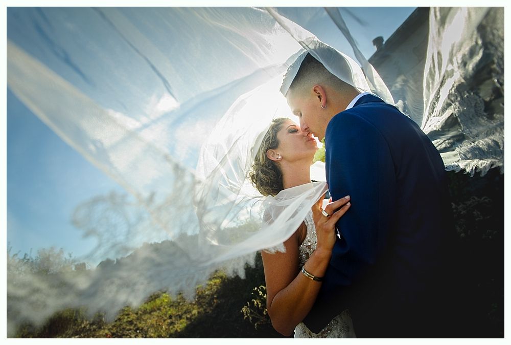 Bride and groom kissing, veil blowing in the wind, with sunlight and mountain backdrop.