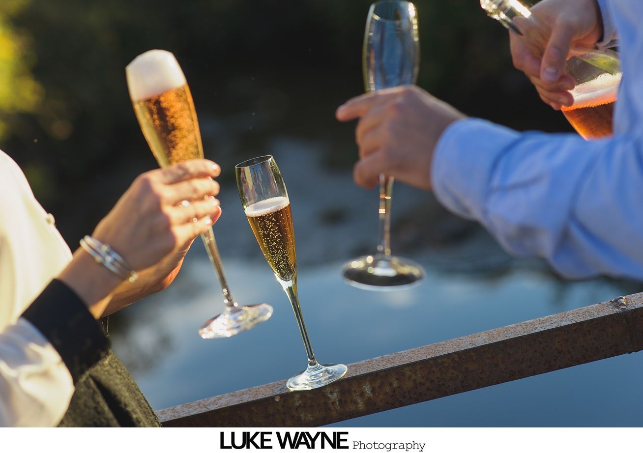 Man hugging woman from behind on a beach; woman smiles, ring visible, blue shirt.