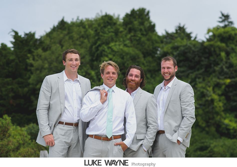 Four men in light gray suits pose in front of a green treeline, one holding a jacket over his shoulder.