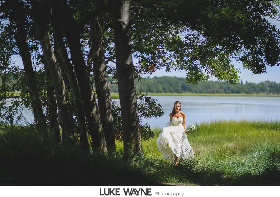 Bride in white gown walks by trees towards a lake on a sunny day.