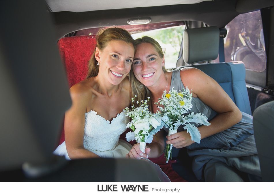 Two smiling women in a car, one in a wedding dress, both holding bouquets.