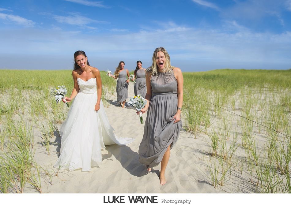 Bride and bridesmaids on a sandy path, laughing, with beach grass and blue sky.