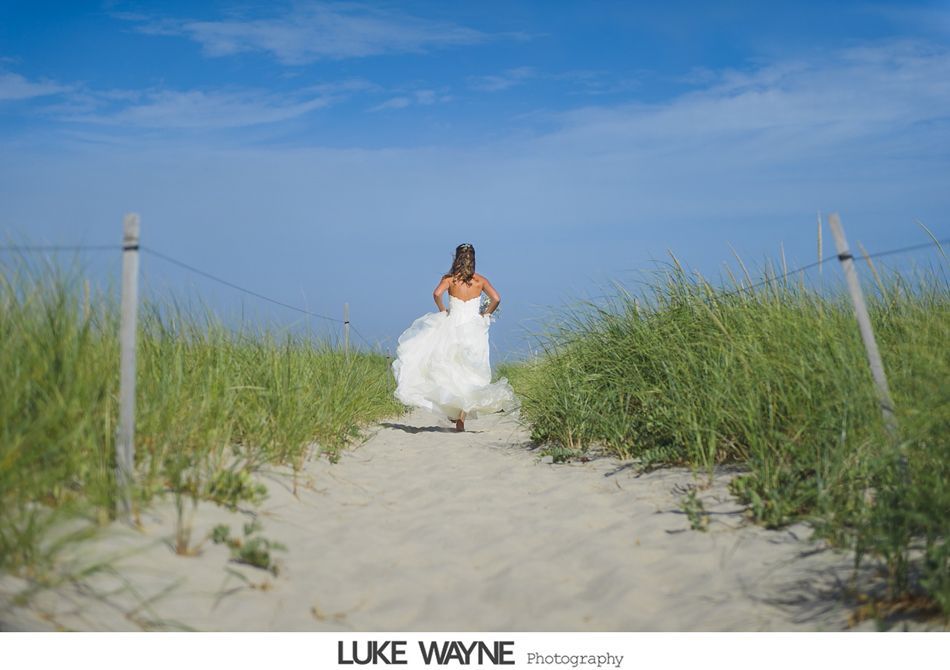 Bride runs down sandy path between tall grass towards the ocean under a blue sky.