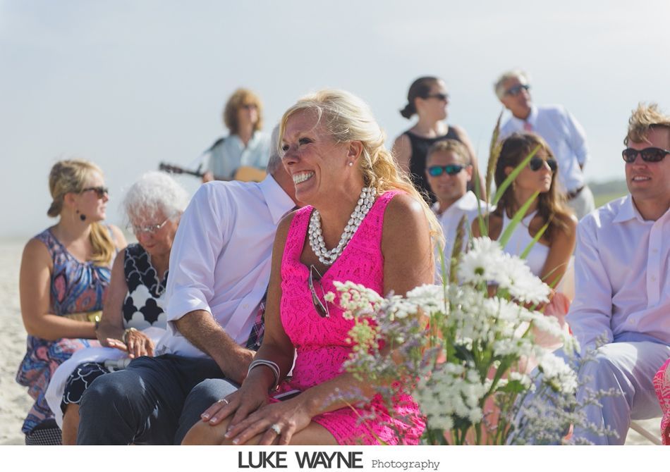 Woman in a pink dress laughs at an outdoor beach wedding; other guests watch.