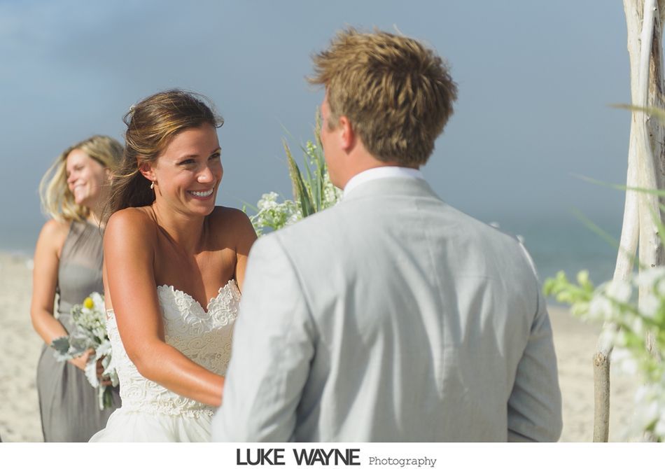 Bride and groom smiling at each other during a beach wedding ceremony. Bridesmaid watches from the side.