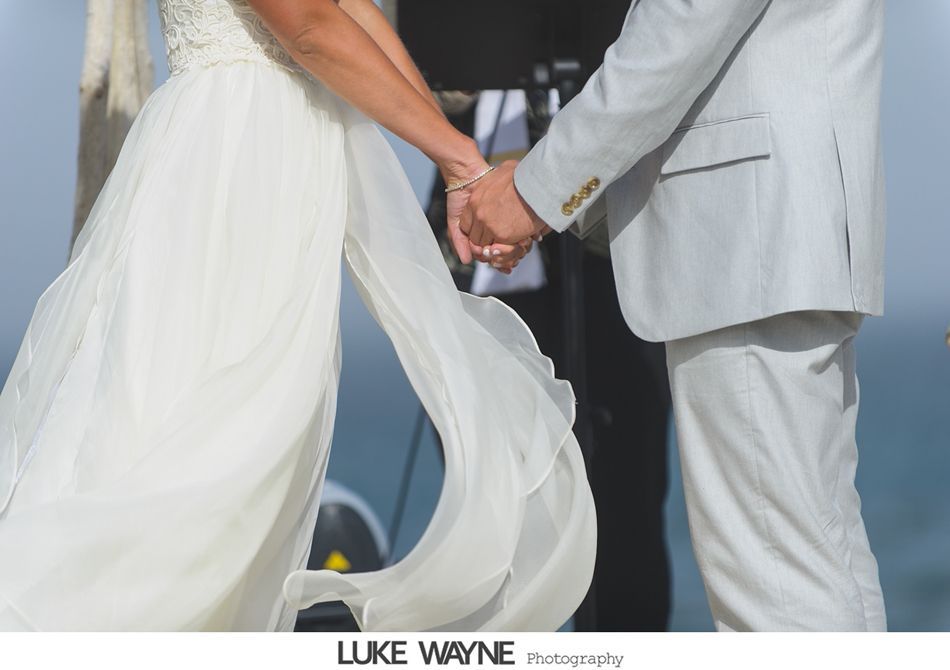 Bride and groom holding hands during a wedding ceremony. White dress, light gray suit, outdoors.
