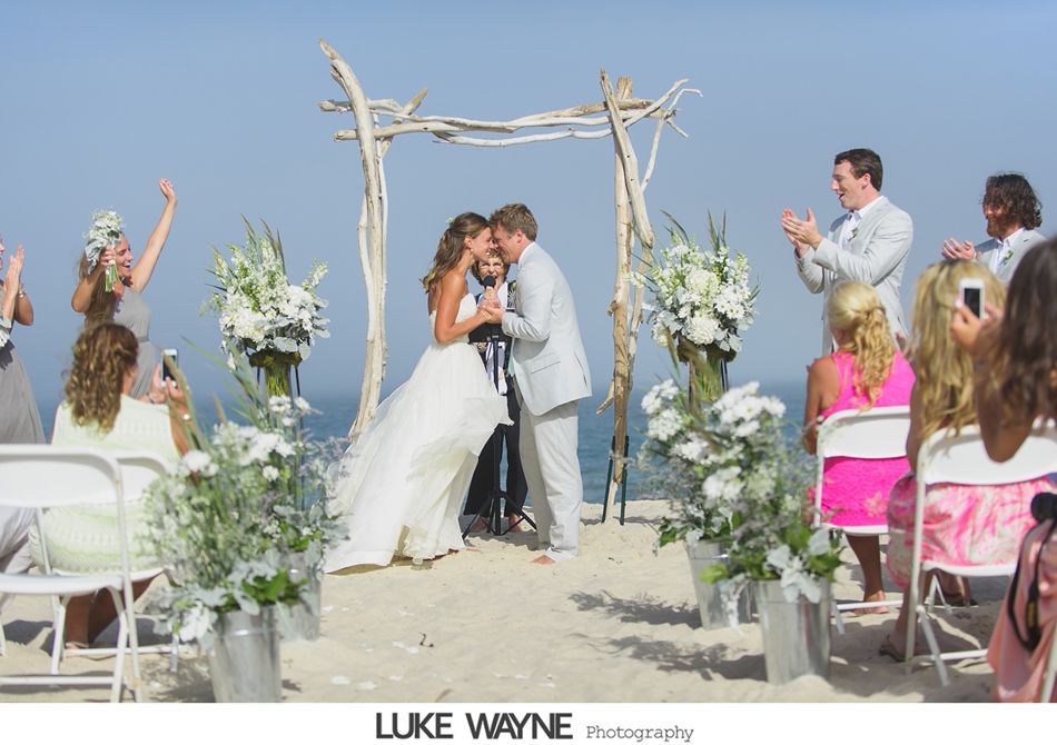 Couple kissing at a beach wedding ceremony, driftwood arch, ocean backdrop, guests clapping.