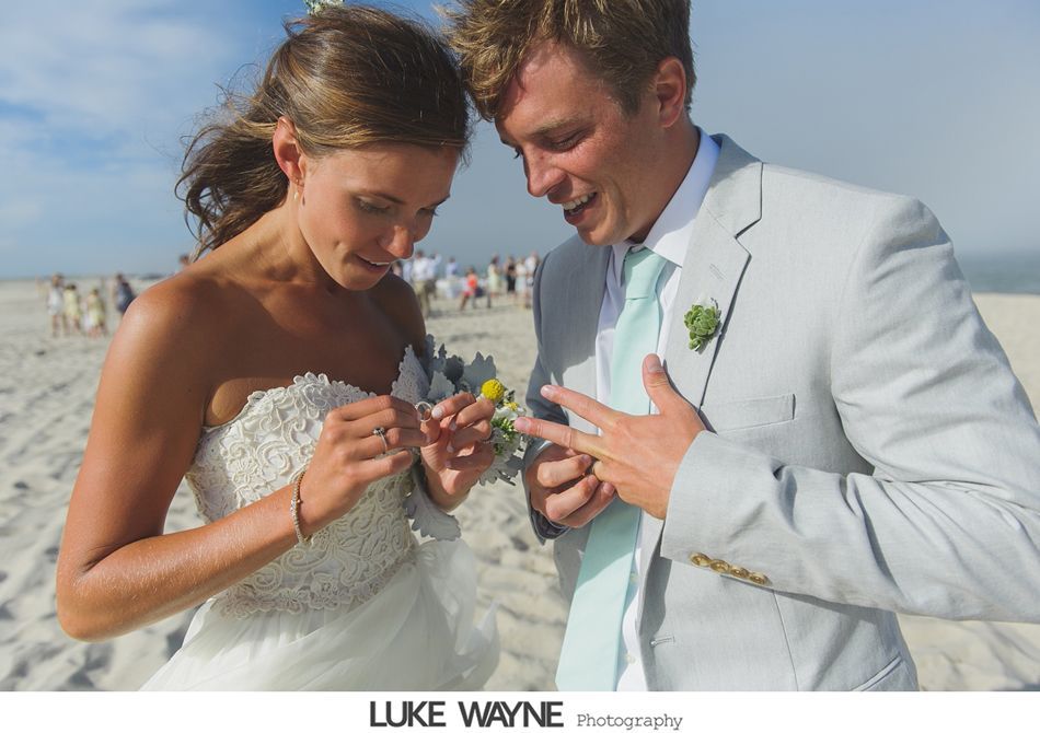 Bride and groom exchange rings on a sunny beach, wearing a white strapless gown and light gray suit.
