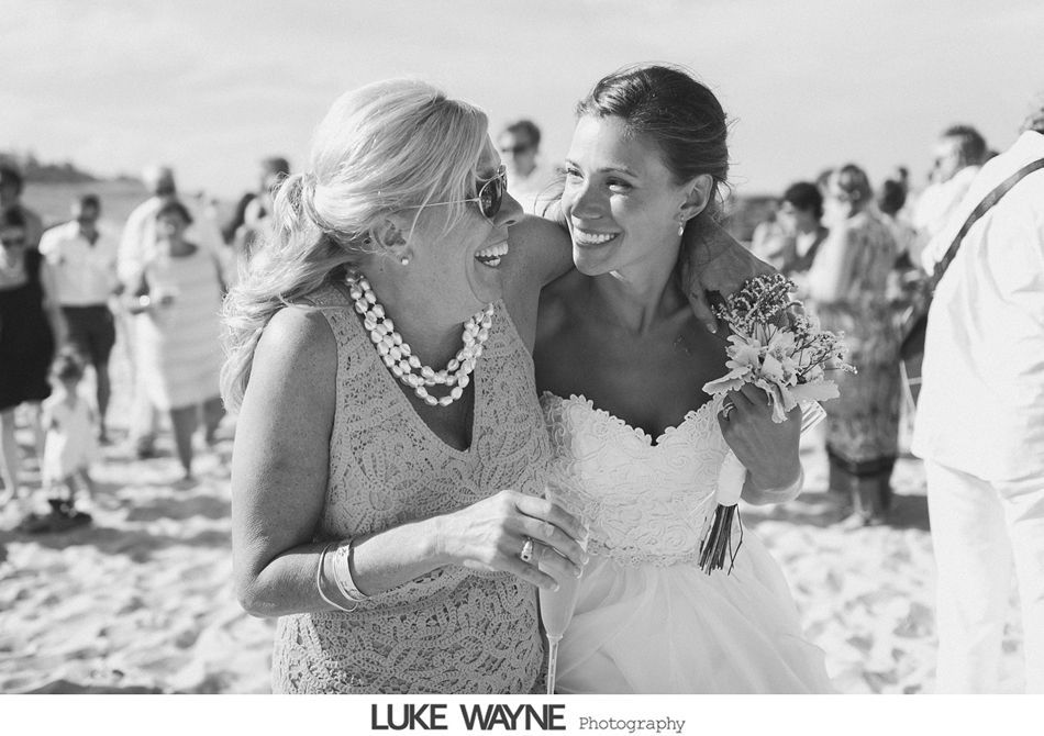 Bride embraces and laughs with older woman at a beach wedding.
