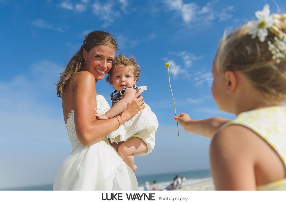 Bride holding baby, facing camera, with child offering flower on beach. Blue sky.