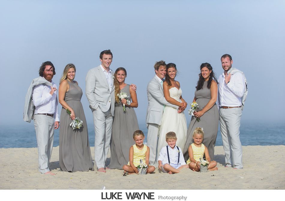 Wedding party on a beach. Bride and groom embrace, bridesmaids in gray gowns, groomsmen in gray suits. Children sit in front.