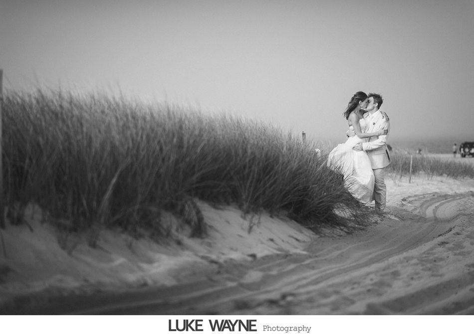 Couple kissing on a beach. Woman in a dress, man in a suit, embrace near dunes. Black and white photo.